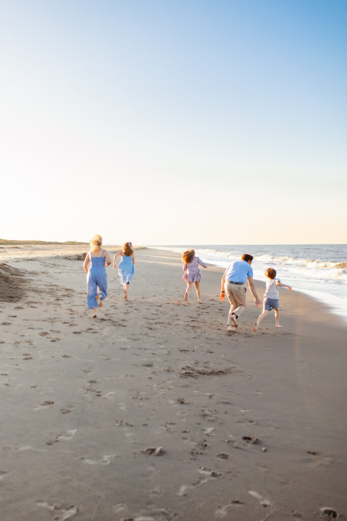 family playing joyfully together running away from the camera chasing each other right at the shore break on a sun drenched play filled family photography session in virginia beach.