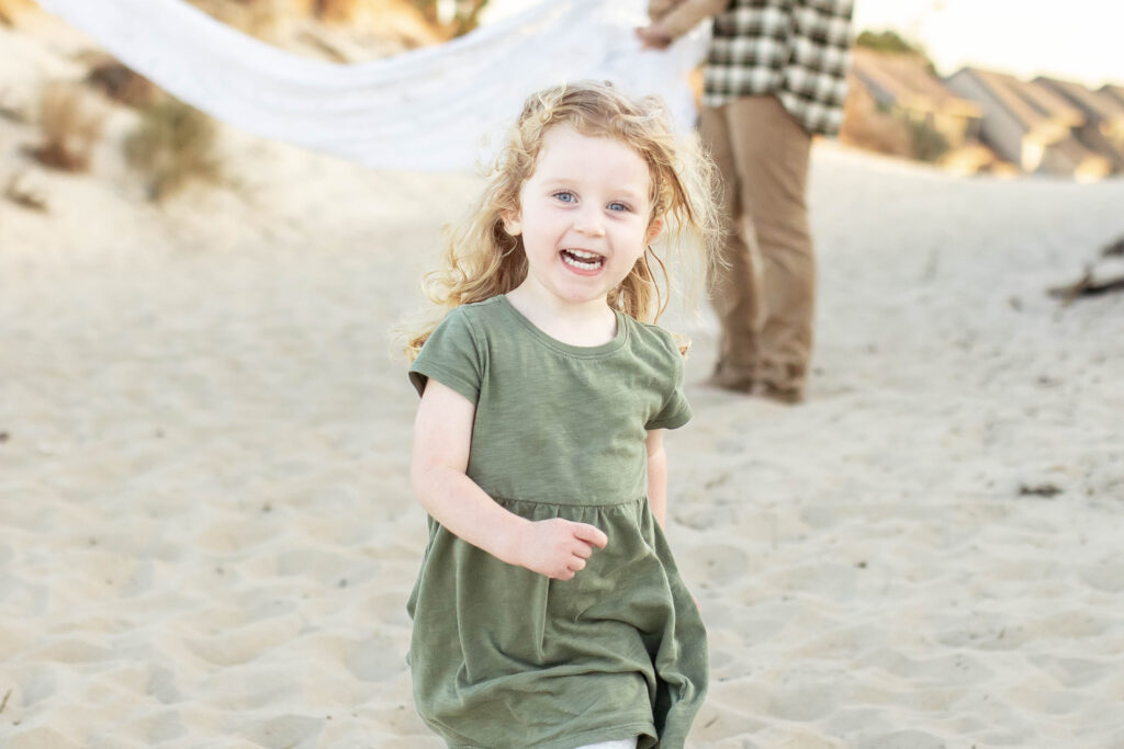 little girl running wild and free dressed in a neutral green on the sand drenched in sunlight during a family portrait session