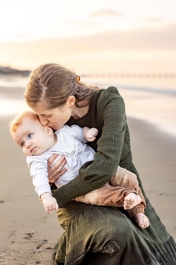 quiet cuddle moment at sunset between mom and baby dressed in neutral greens and whites on the bay with the calm water in the background with the sun setting behind them.  