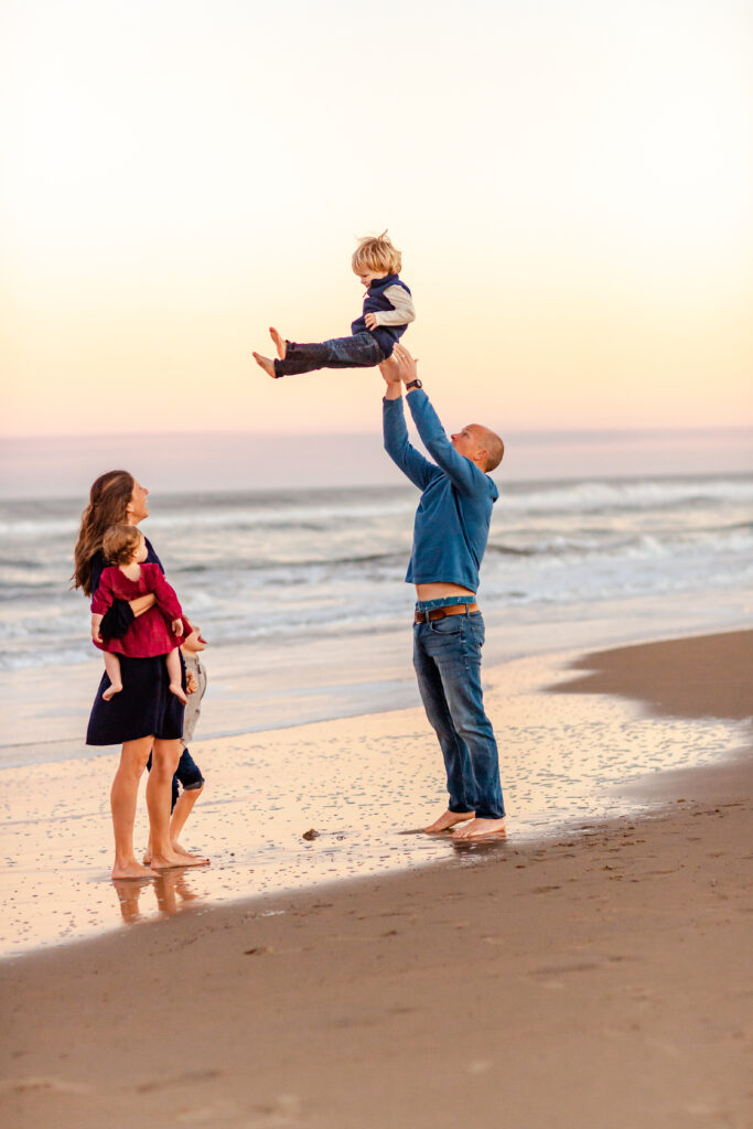 family of four dressed in navy and wine play together as dad tosses his youngest son in the air during a water color sunset during a playful family session