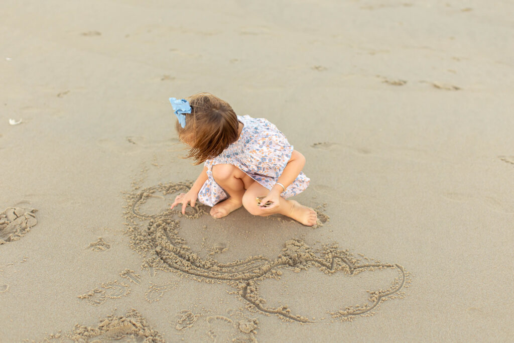 little girl with blue bow and pink flowered dress kneels down in the sand during sunset to draw a rainbow with her fingers in the shorebreak at a family session in virginia beach, virginia