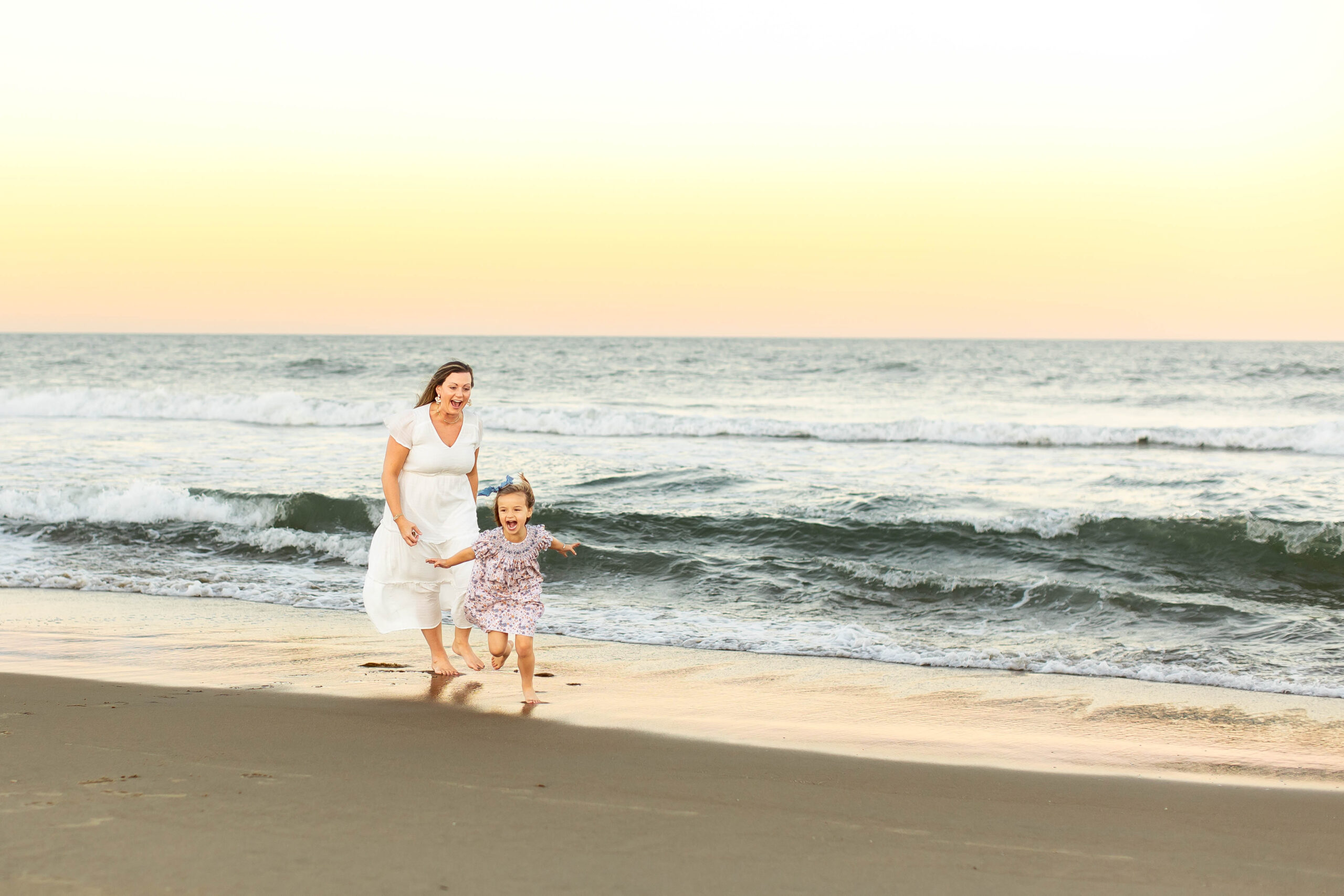 beautiful mother and daughter run in the shore break at the virginia beach ocean front laughing and giggling during a water color sunset during a playful family photoshoot