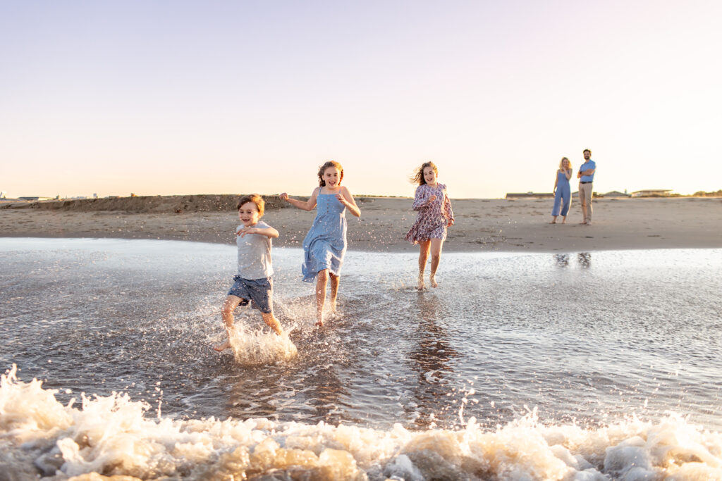 Three Chilfren laugh and run through the ocean surf during a candid family photo session at the beach with Brooke Tucker Photography, capturing joy and chaos of real family connection in Coastal Virginia
