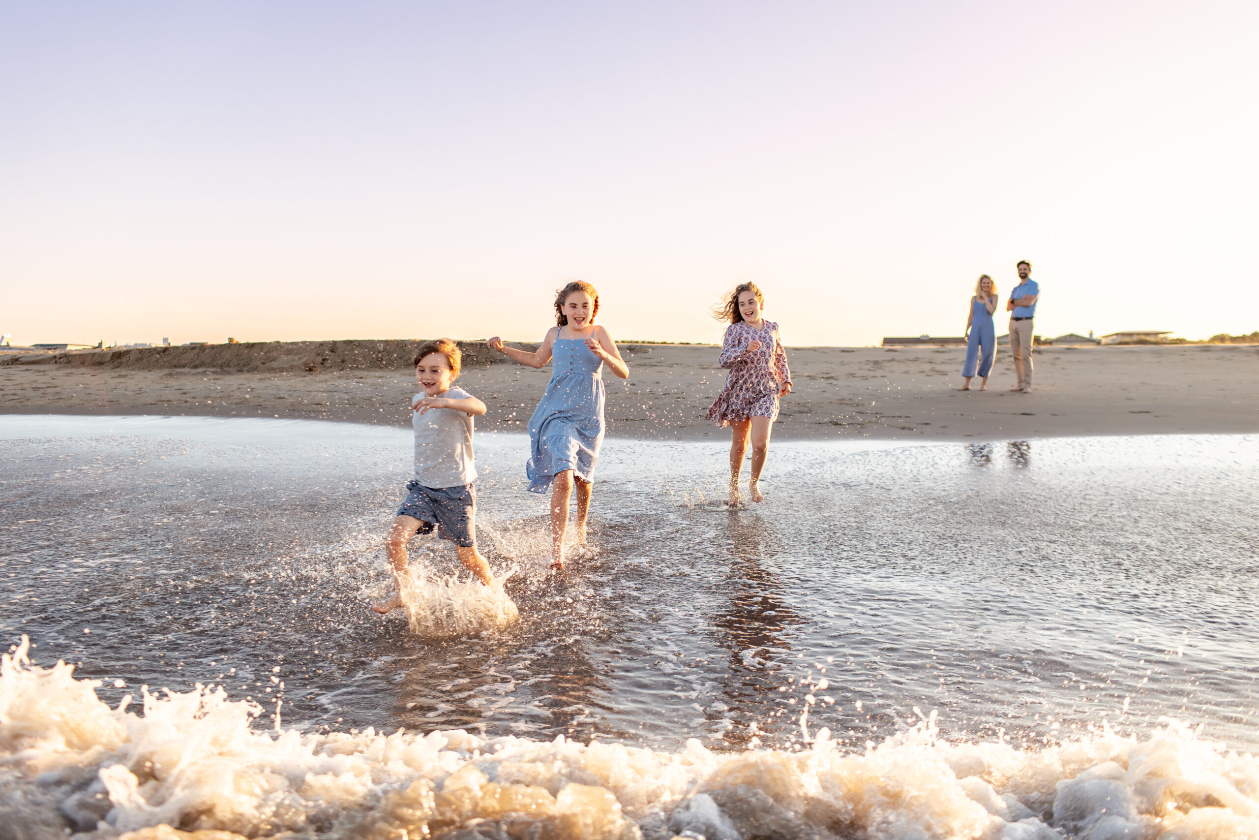 Three Chilfren laugh and run through the ocean surf during a candid family photo session at the beach with Brooke Tucker Photography, capturing joy and chaos of real family connection in Coastal Virginia