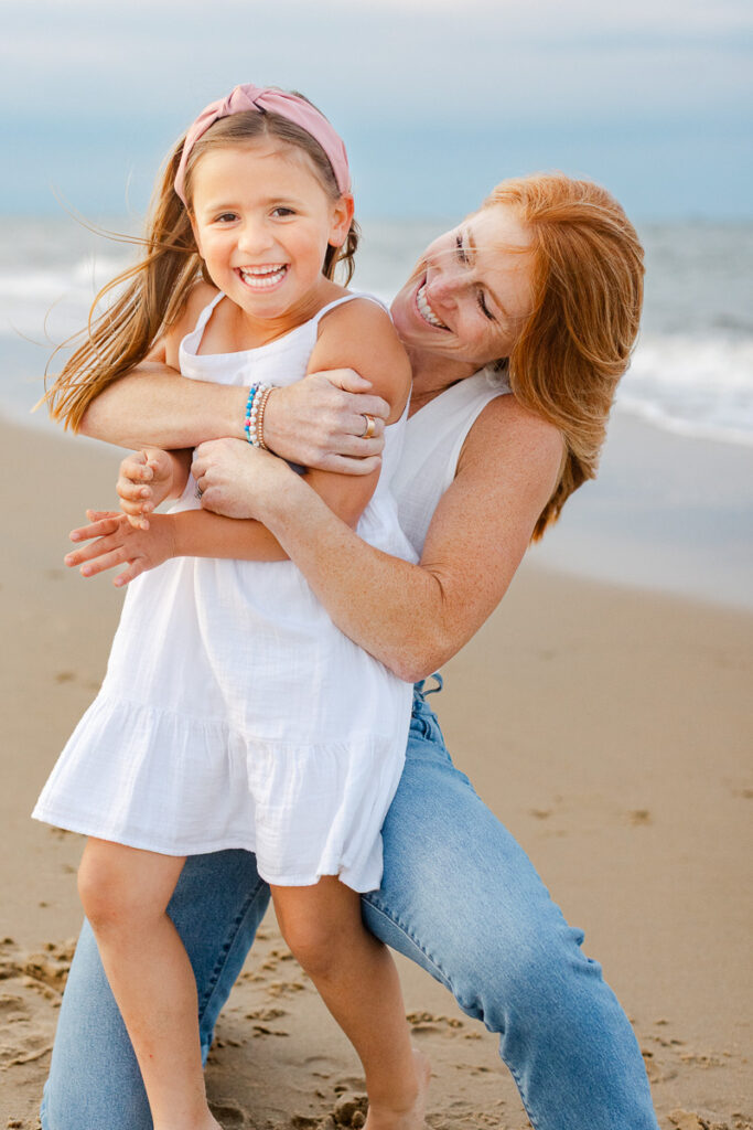 A other wraps her arms around her smiling daughter from behind, both looking out toward the beach with joy
